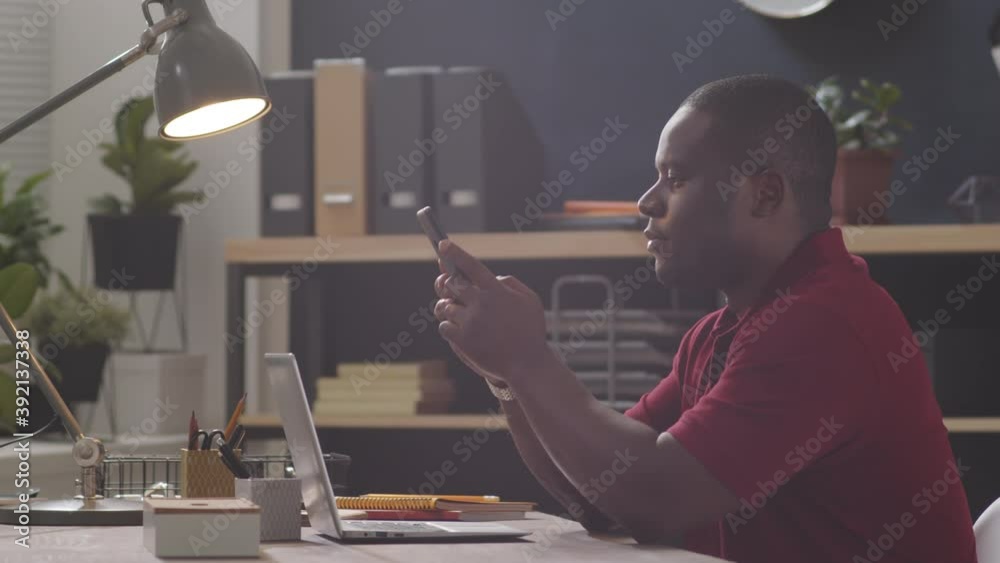 Side view tracking shot of black businessman surfing the mobile Internet on smartphone while sitting at office desk Side view tracking shot of black businessman surfing the mobile Internet on smartphone while sitting at office desk