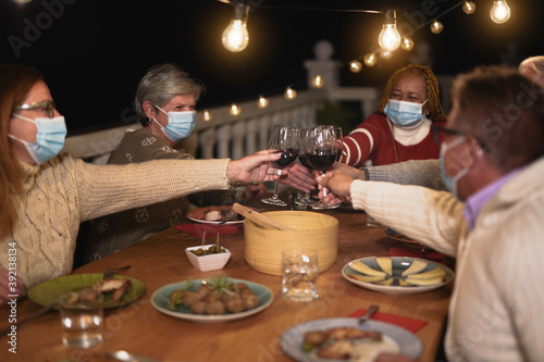 Multiracial senior people celebrate christmas together with dinner outdoor while wearing surgical face mask - Friends cheering with red wine photo