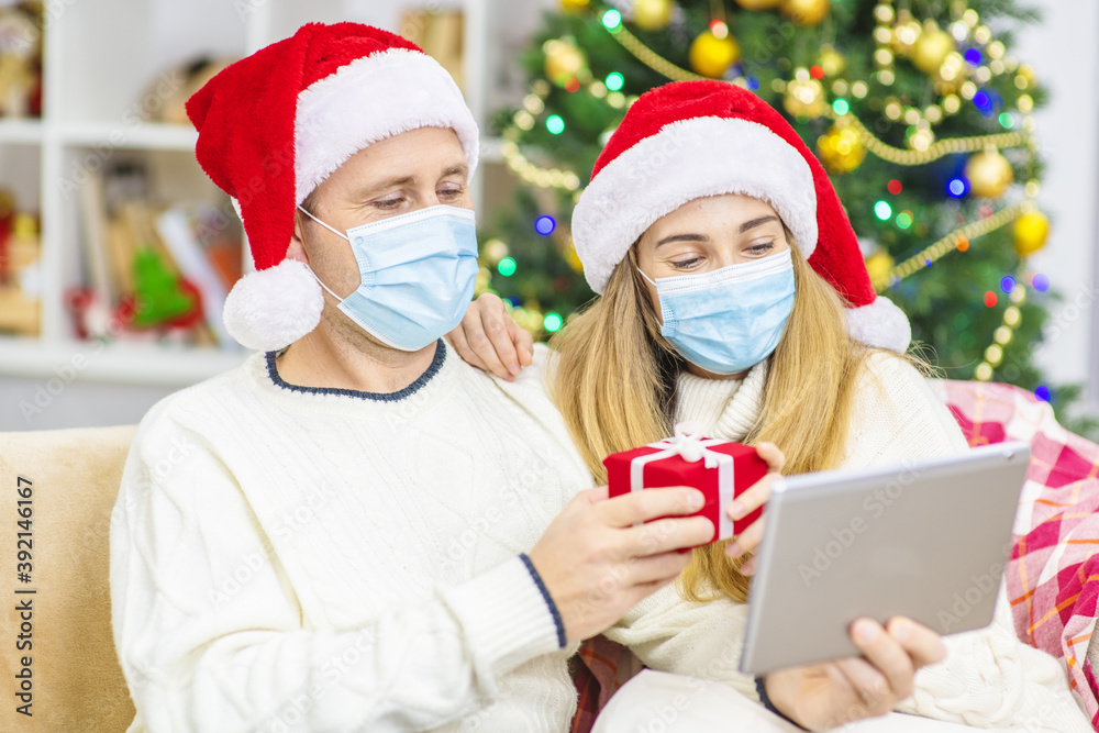 A young married couple with medical masks are sitting on the sofa in Santa hats against the background of a Christmas tree and congratulating relatives with Christmas (New Year) online using a tablet. A young married couple with medical masks are sitting on the sofa in Santa hats against the background of a Christmas tree and congratulating relatives with Christmas (New Year) online using a tablet.