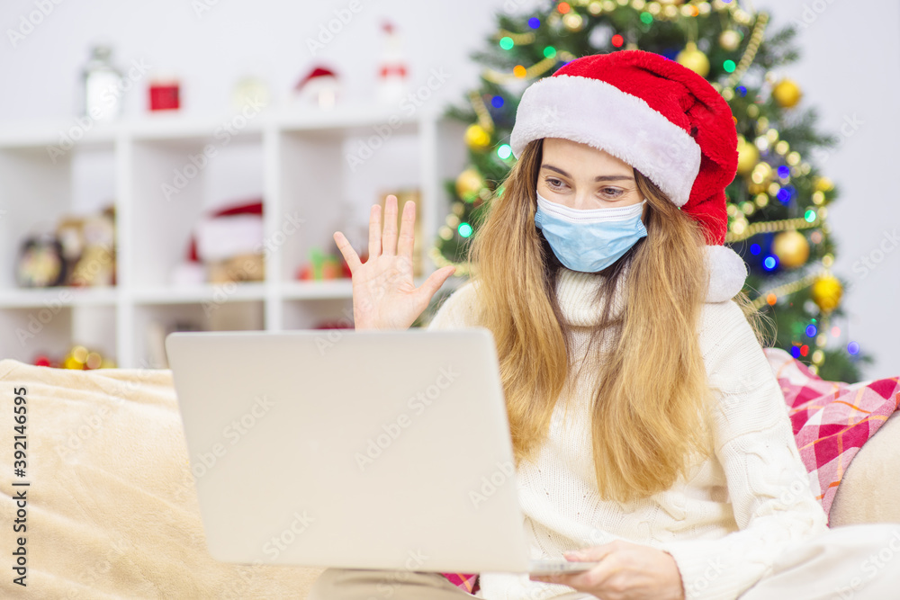 A young girl with a medical mask and a santa hat waves a welcome hand during an online call for christmas (new year) to relatives. Coronavirus pandemic during the winter holidays. Lockdown at Christma A young girl with a medical mask and a santa hat waves a welcome hand during an online call for christmas (new year) to relatives. Coronavirus pandemic during the winter holidays. Lockdown at Christma