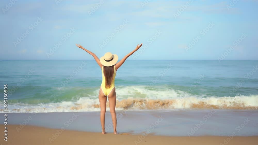 A young woman in a one-piece bathing suit with her back to the camera greets the incoming ocean tide and waves with joy and extended arms. copy space A young woman in a one-piece bathing suit with her back to the camera greets the incoming ocean tide and waves with joy and extended arms. copy space