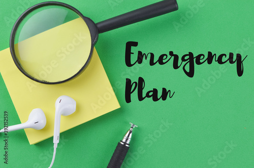 Selective focus of magnifying glass, memo note, earphones and pen over a green bckground written with text EMERGENCY PLAN. 