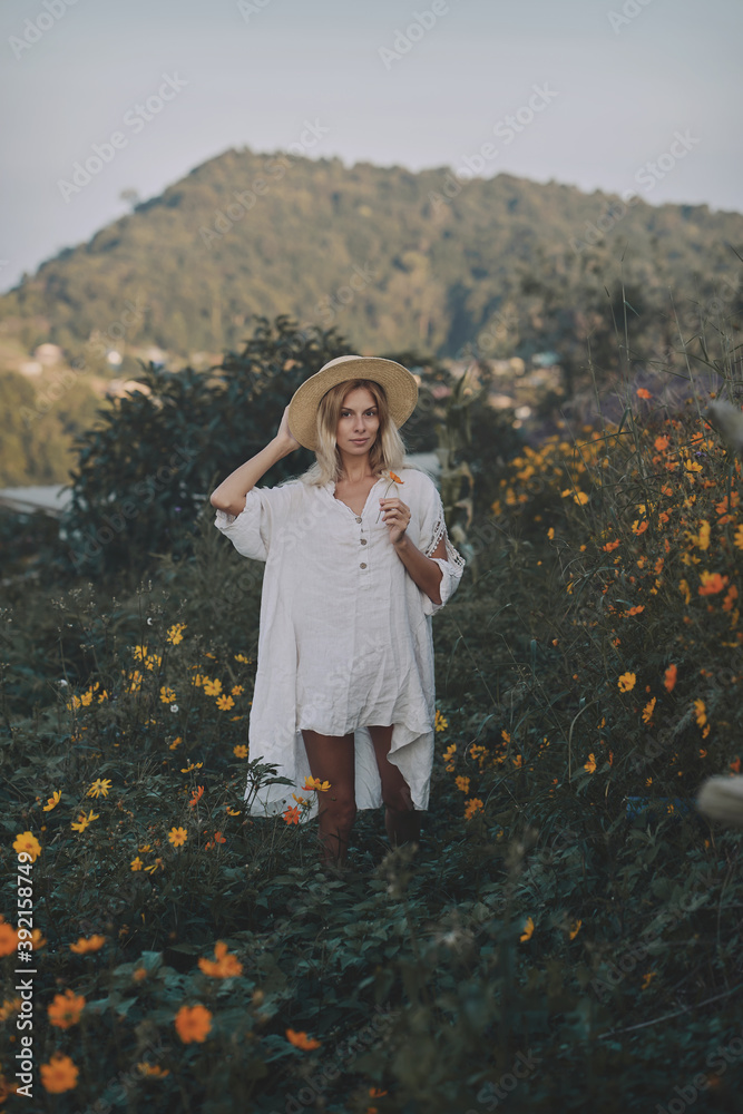 The blonde in a linen dress and straw hat walks among herbs and wildflowers on a meadow in the mountains. A woman in the Boho style, resting in the countryside, a simple village lifestyle