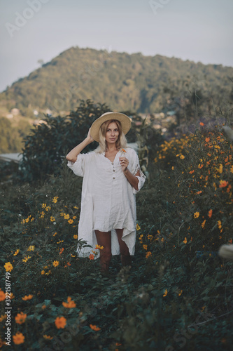 Obraz na płótnie The blonde in a linen dress and straw hat walks among herbs and wildflowers on a meadow in the mountains. A woman in the Boho style, resting in the countryside, a simple village lifestyle w salonie
