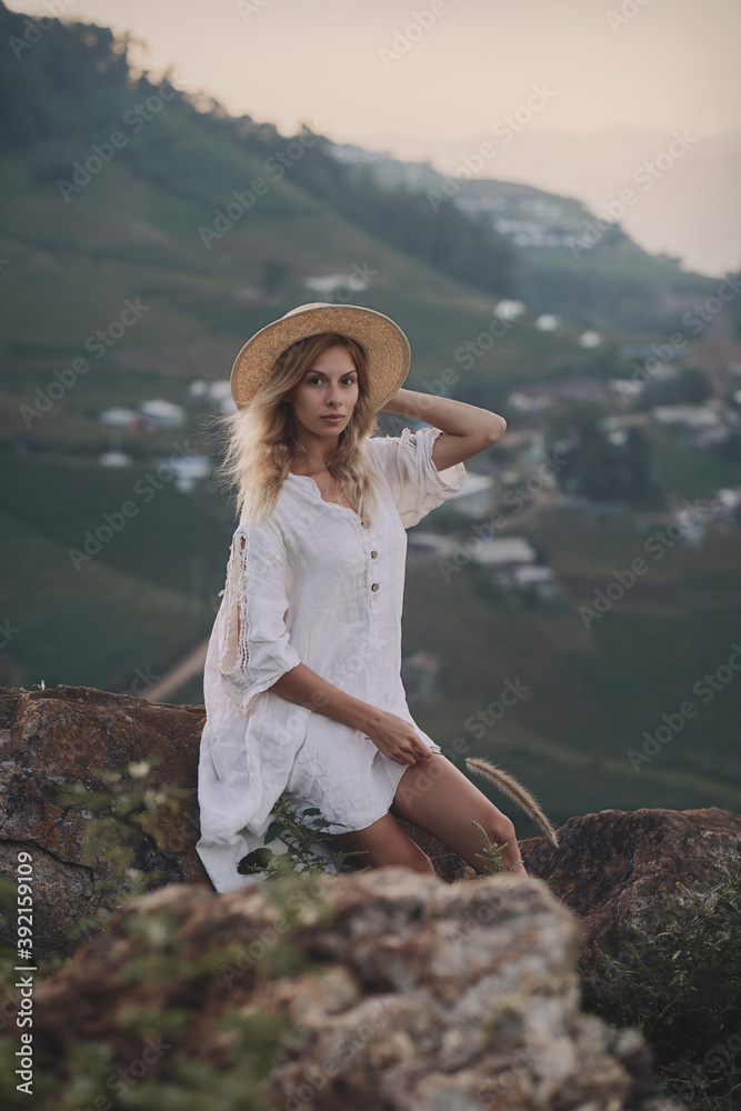 The blonde in a linen dress and straw hat sits on a viewpoint in the mountains. A woman in the Boho style, resting in the countryside, a simple village lifestyle