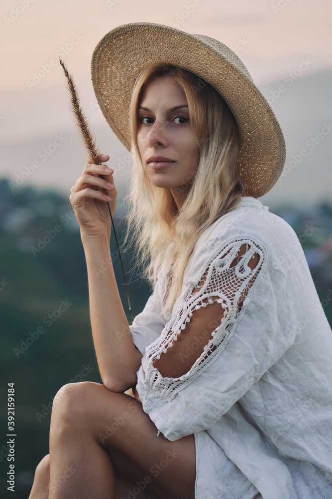 The blonde in a linen dress and straw hat sits on a viewpoint in the mountains. A woman in the Boho style, resting in the countryside, a simple village lifestyle