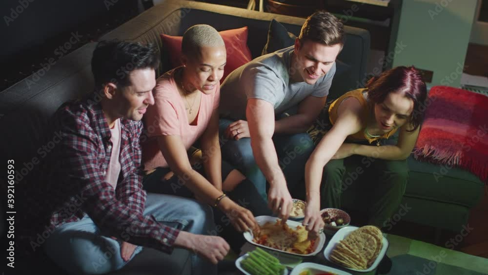 Group of friends sitting on sofa at home eating snacks and laughing watching evening comedy TV together - shot in slow motion