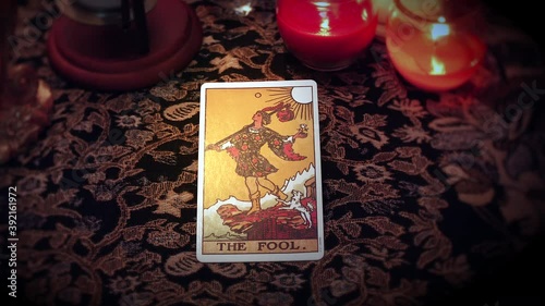 A mystical fortune teller selects a Tarot card and places it on the enchanted table. The fool card is drawn and displayed in front of the candles and voodoo ornaments.