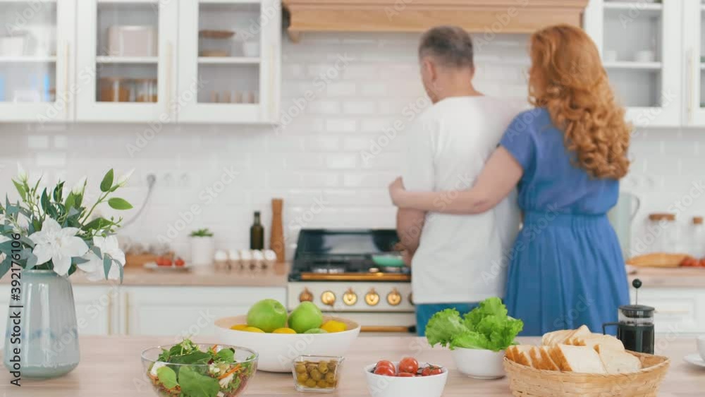 Rear view of middle-aged beautiful Caucasian woman with long curly hair embracing her handsome husband standing at stove in the kitchen and cooking Rear view of middle-aged beautiful Caucasian woman with long curly hair embracing her handsome husband standing at stove in the kitchen and cooking