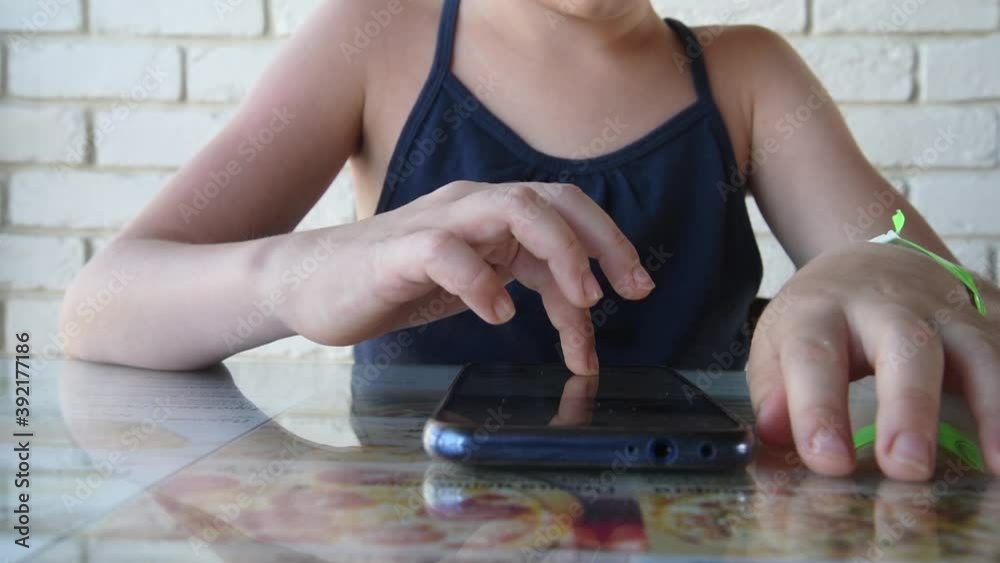 The mobile phone is on the table in front of the child. The girl is playing on her phone. The child is passionate about the game. Hands close-up The mobile phone is on the table in front of the child. The girl is playing on her phone. The child is passionate about the game. Hands close-up