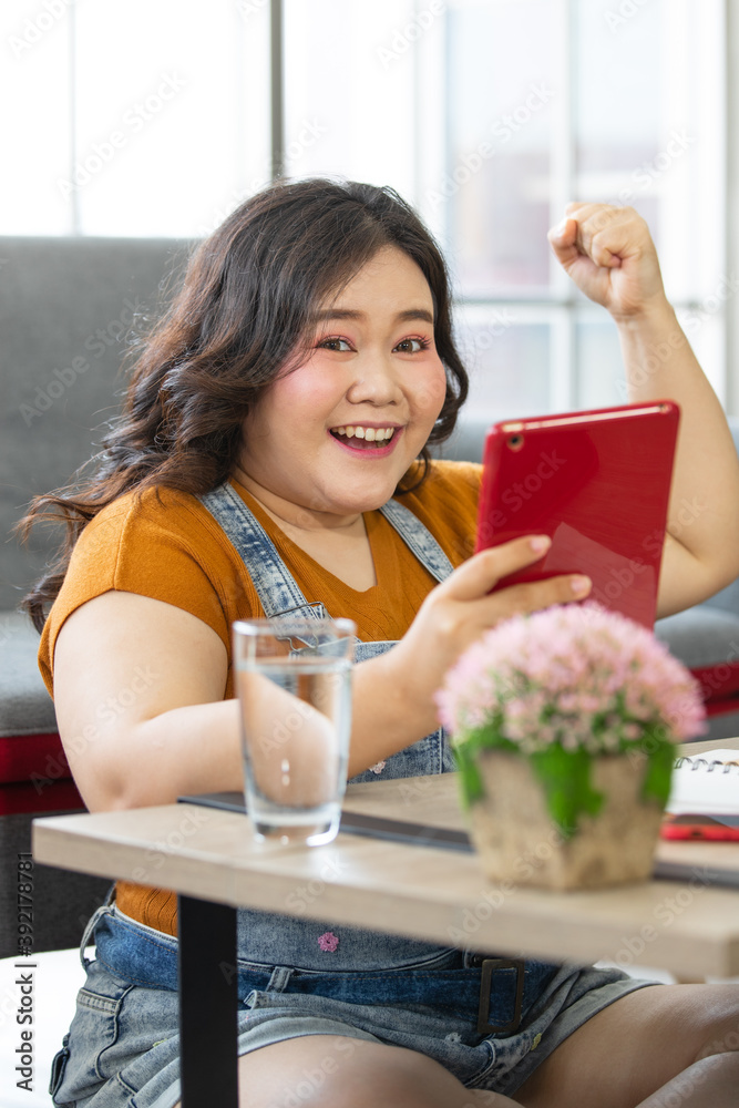 portrait of fat asian woman wearing casual cloth sitting on the floor in a house holding a tablet and hand up looking at a camera with smiling face. Selective focus on a happy lady face