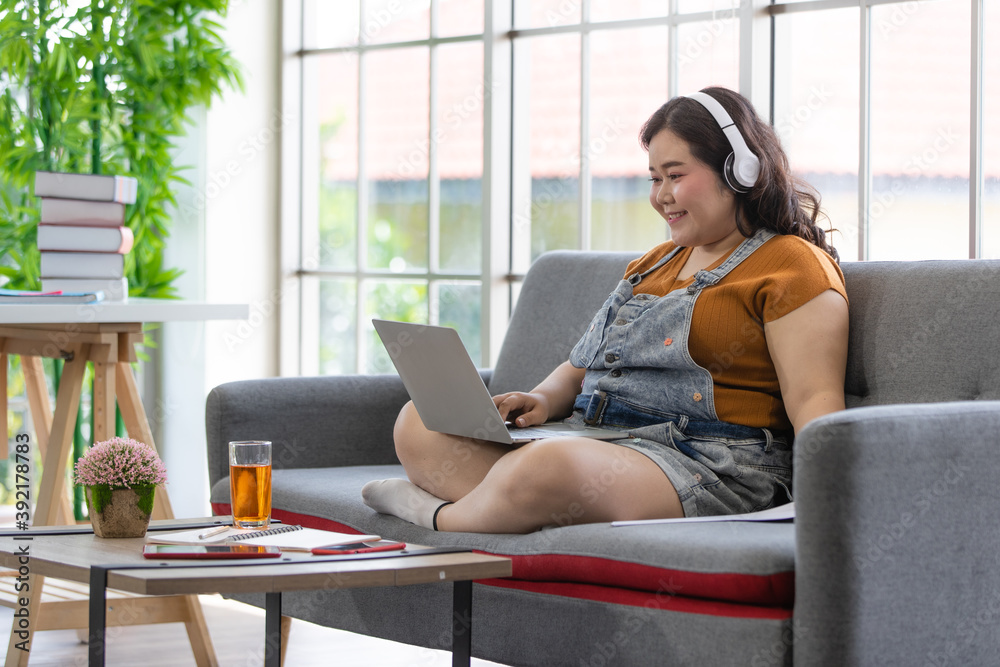 © Bangkok Click Studio - fat female wearing casual cloth with headphones smiling and sitting on a couch looking at laptop computer on her lap during doing online communication in a house. Study and work from home concept