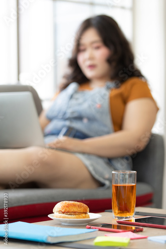 hamburger in a white plate and glass of juice on a table with a blur background of fat woman sitting on a couch at home looking to food. Selective focus on hamburger