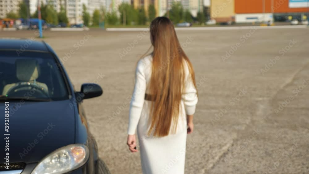 Blurred elegant businesswoman in long dress of white colour opens black car and sits on driver seat in automobile on large parking lot in summer Blurred elegant businesswoman in long dress of white colour opens black car and sits on driver seat in automobile on large parking lot in summer
