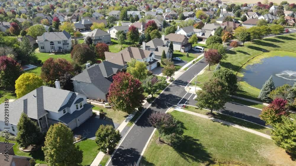 Overhead aerial view of colorful autumn trees, residential houses and yards with drainage pond along suburban street in Chicago area. Midwest USA. 4K
