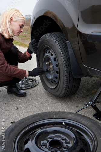 A young blonde woman removes the wheel with a key near her car with a flat tire. © Pavlo