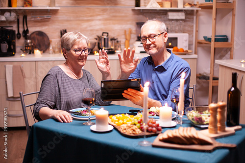 Mature man and woman waving at tablet pc during video conference in kitchen celebrating relationship. Couple sitting at the table, browsing, talking, using internet, celebrating their anniversary in