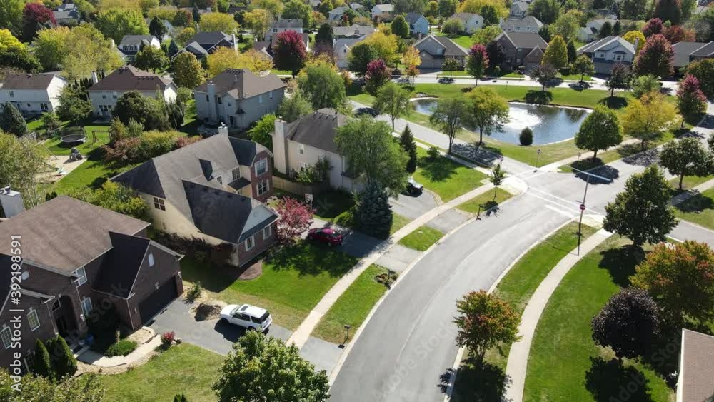 Overhead aerial view of colorful autumn trees, residential houses and yards with drainage pond along suburban street in Chicago area. Midwest USA. 4K

