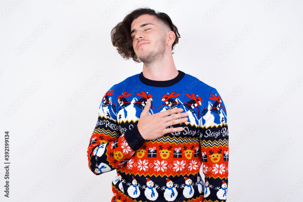 Satisfied smiling Young handsome Caucasian man wearing Christmas sweater, keeps hands on belly, being in good mood after eating delicious supper, demonstrates she is full. Pleasant feeling in stomach.