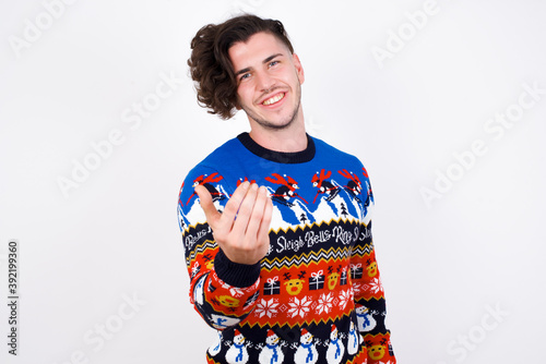 Young handsome Caucasian man wearing Christmas sweater against white wall, inviting you to come, confident and smiling making a gesture with hand, being positive and friendly.