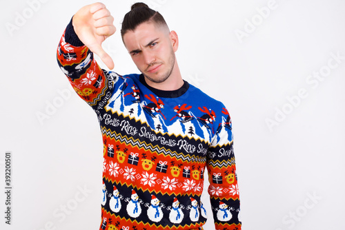 Foto Discontent Young handsome Caucasian man wearing Christmas sweater against white wall shows disapproval sign, keeps thumb down, expresses dislike, frowns face in discontent