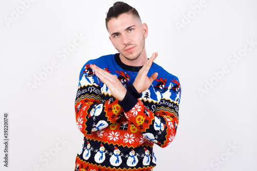 Young handsome Caucasian man wearing Christmas sweater against white wall Has rejection angry expression on face and crossing hands doing refusal negative sign.