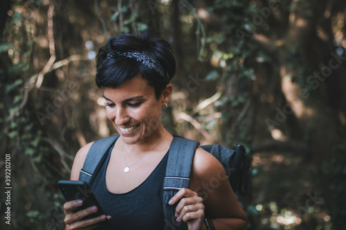 Woman looking at her phone smiling in the forest while hiking wearing a backpack