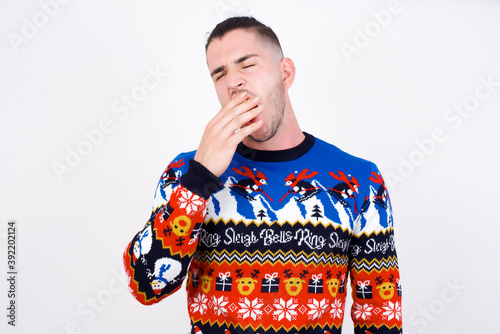 Sleepy Young handsome Caucasian man wearing Christmas sweater against white wall yawning with messy hair, feeling tired after sleepless night, yawning, covering mouth with palm.