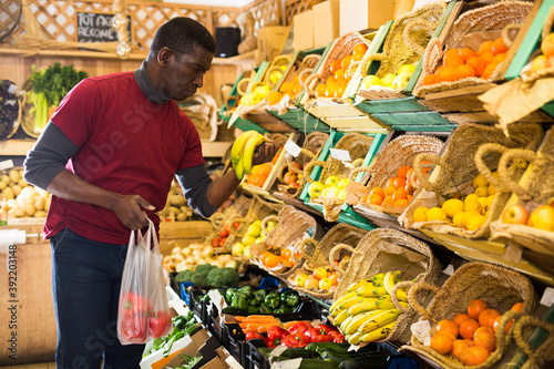 Interested African American man making purchases in greengrocery store, looking for fresh vegetables and fruits..