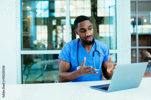 Portrait of a male doctor wearing blue scrubs uniform using laptop to talk to patient online © Carlos David