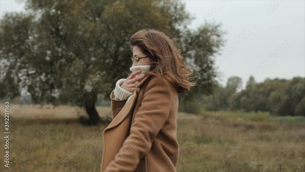 An energetic young girl in glasses stands against a tree background and laughs while wrapping herself in a warm white sweater and coat An energetic young girl in glasses stands against a tree background and laughs while wrapping herself in a warm white sweater and coat