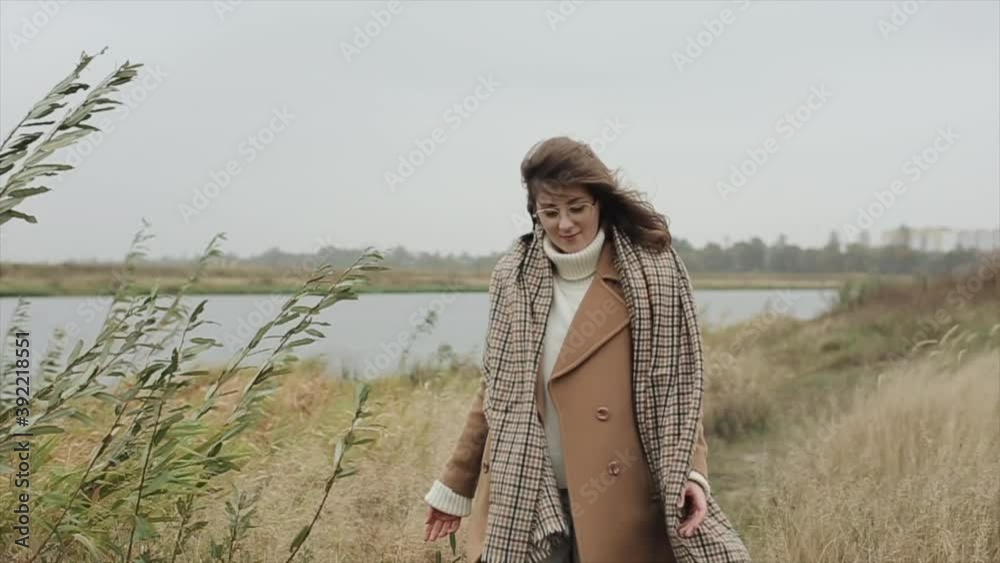 Beautiful young woman goes along the river bank in a coat and a checkered scarf and touches the grass with her hands Beautiful young woman goes along the river bank in a coat and a checkered scarf and touches the grass with her hands