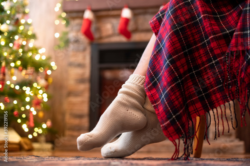 Bottom view of woman legs in knitted winter socks sitting on armchair covered plaid near christmas tree and fireplace. Bottom view
