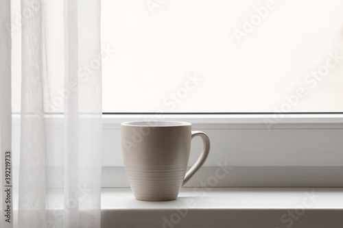 Big brown mug on window sill at curtains. Waking with fresh coffee in morning concept. Closeup. Front view. Empty place for text on white glass.