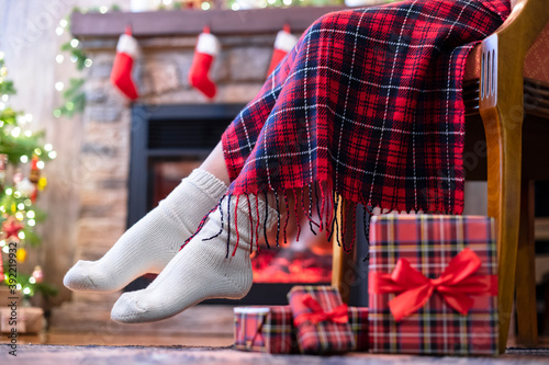 Woman legs in a winter socks covered plaid sitting and relaxation on armchair near fireplace and christmas tree pakking gift boxes for family. Bottom view.