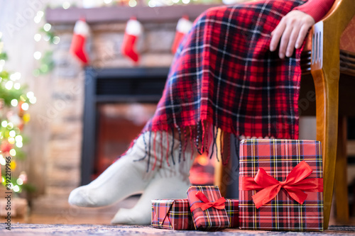 Woman legs in a winter socks covered plaid sitting and relaxation on armchair near fireplace and christmas tree pakking gift boxes for family. Bottom view. photo