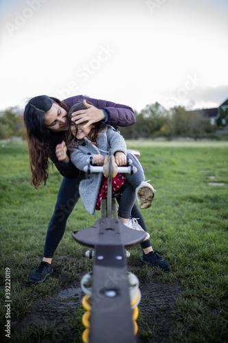 Happy mother playfully covers the eyes of her little daughter on a seesaw