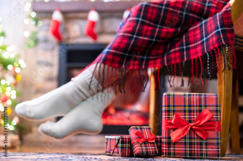 Woman legs in a winter socks covered plaid sitting and relaxation on armchair near fireplace and christmas tree pakking gift boxes for family. Bottom view.