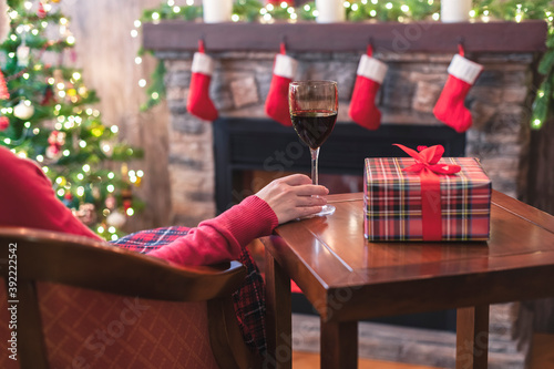 Happy woman covered plaid with glass of wine sitting and relaxation on armchair near fareplace and fir-tree after finishing pakking gift boxes for family. photo