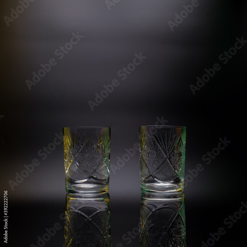 Two crystal glasses on a black background. Decorated with carvings, ornaments. On the edges there are multi-colored highlights. Reflection.