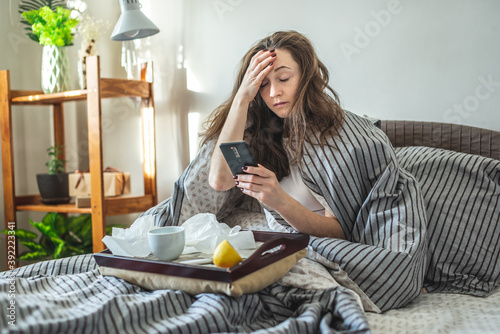 Young sick woman is sitting in bed wrapped in a blanket, holding her aching head and using her phone photo