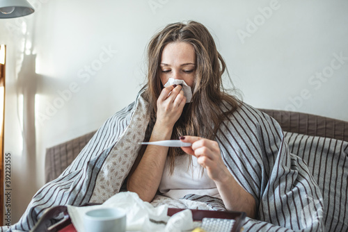 Young sick woman is sitting in bed wrapped in a blanket and using a handkerchief and looking at the thermometer photo