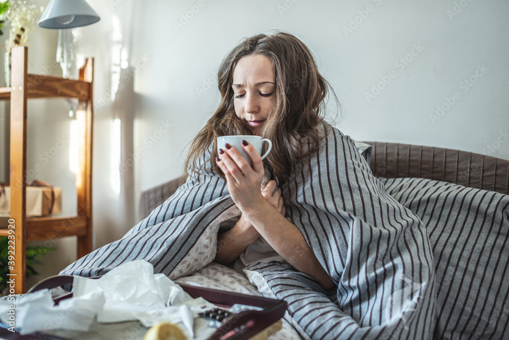Young sick woman is sitting in bed wrapped in a blanket and drinking beverage. Concept of illness and cure at home Young sick woman is sitting in bed wrapped in a blanket and drinking beverage. Concept of illness and cure at home