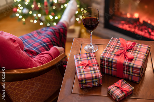 Alone woman covered plaid with glass of wine sitting and relaxation on armchair near fareplace and christmas tree after finishing pakking gift boxes for family. photo