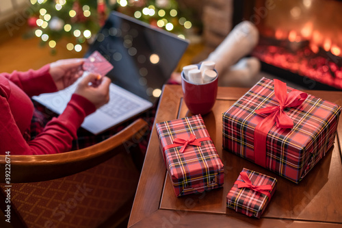 Woman holding credit card using laptop for making order sitting at table with packaging gift, cup of cocoa and marshmallows near fireplace and christmas tree. Online shopping concept