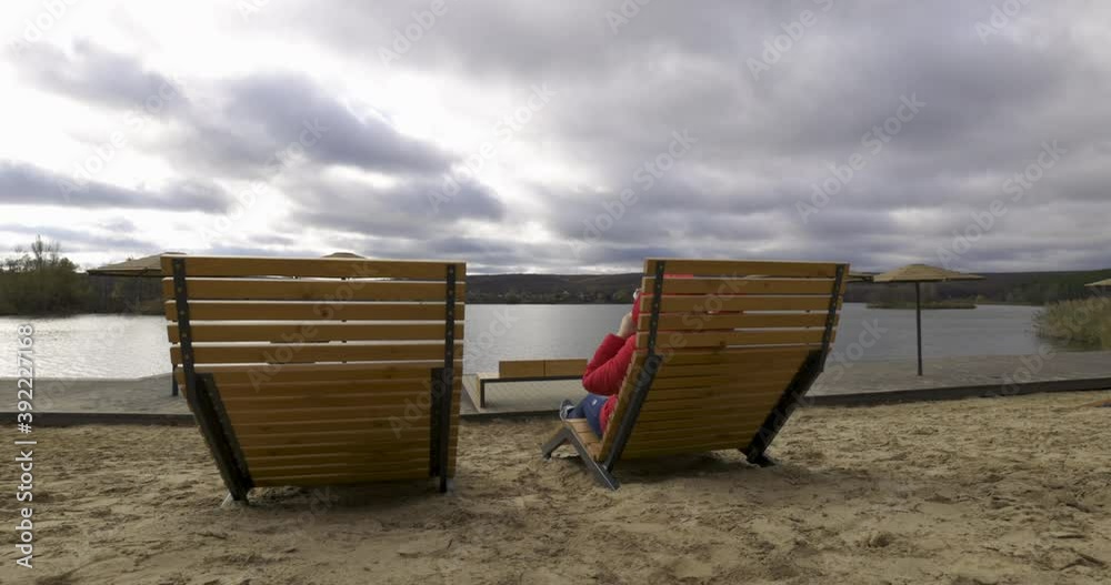 Young woman drinks warming hot coffee or tea from a thermos in cold weather, while sitting on the deckchair on the beach. Happy girl walking on the outdoors at the sea, slow motion 60 fps. Relaxation. Young woman drinks warming hot coffee or tea from a thermos in cold weather, while sitting on the deckchair on the beach. Happy girl walking on the outdoors at the sea, slow motion 60 fps. Relaxation.