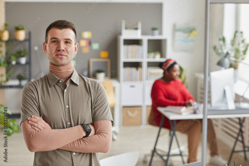 Obraz premium Portrait of young business leader standing with arms crossed and looking at camera at office with woman working in the background