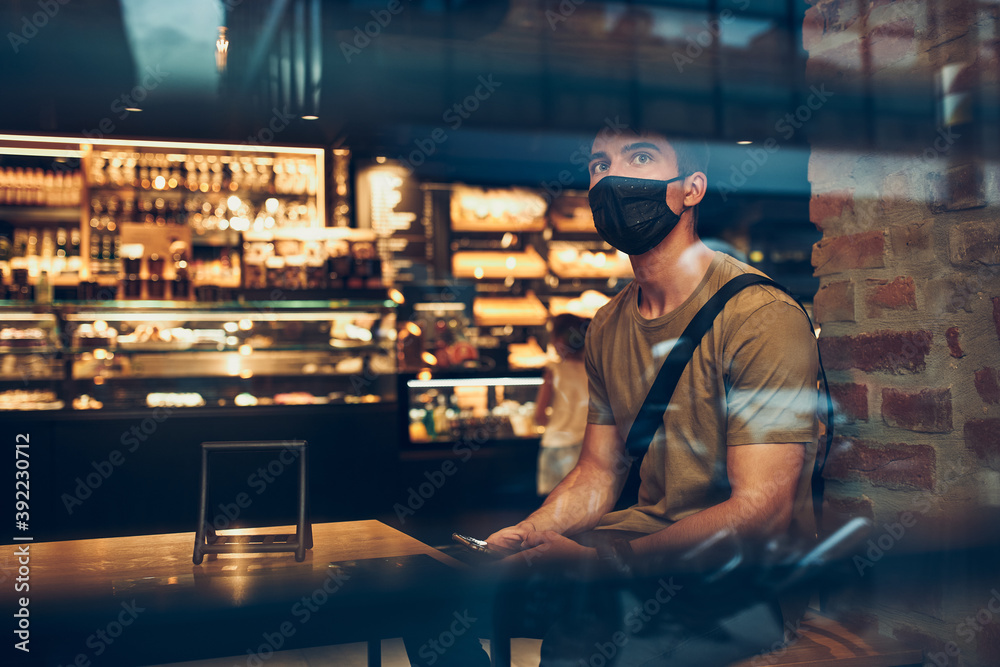 Young man sitting in coffee shop at store front in the city center in the evening, wearing the face mask to avoid virus infection and to prevent the spread of disease in time of coronavirus Young man sitting in coffee shop at store front in the city center in the evening, wearing the face mask to avoid virus infection and to prevent the spread of disease in time of coronavirus