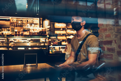 Young man sitting in coffee shop at store front in the city center in the evening, wearing the face mask to avoid virus infection and to prevent the spread of disease in time of coronavirus