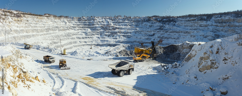 Panorama of a huge snow-covered stone quarry in sunny weather with various mining machines in the foreground. Industrial landscape.